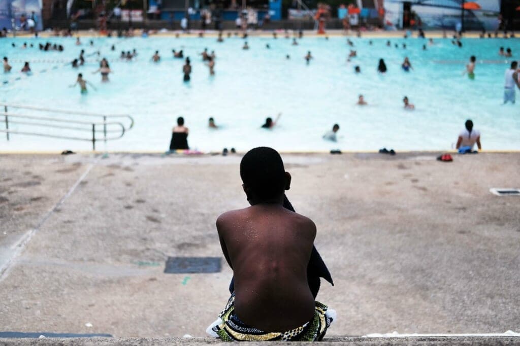 A boy sits on the deck in front of a pool with people swimming