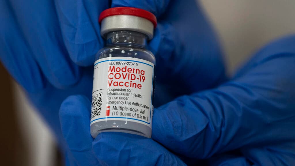 Hands wearing blue medical gloves holds a glass bottle with a red lid labeled "Moderna COVID-19 Vaccine"