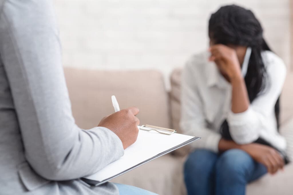A therapist takes notes as a woman sits on a couch holding her head