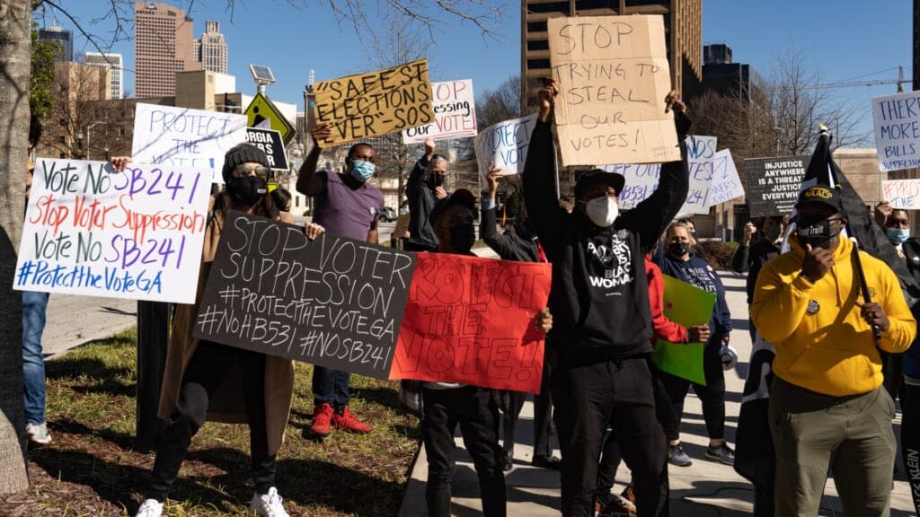 Demonstrators wearing lower-face masks hold signs protesting voting restrictions.