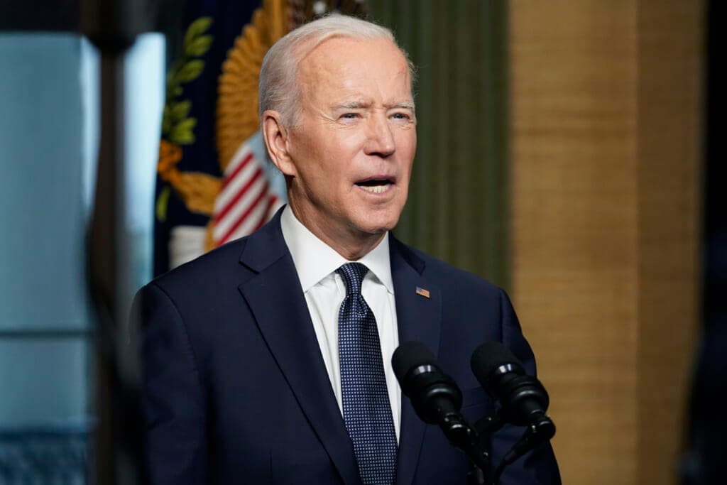 President Joe Biden behind two microphones wearing a navy jacket and blue checked tie