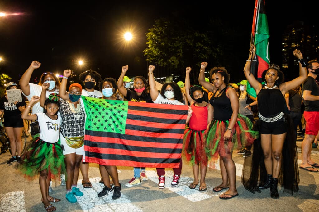 A group of people with a green, black and orange American flag raise their fists in the air