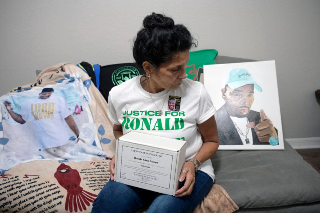 Mona Hardin sits on a bed holding the boxed ashes of her son, Ronald Greene
