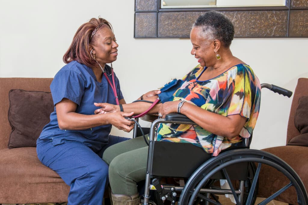 A caregiver in medical scrubs takes the blood pressure of a woman in a wheelchair