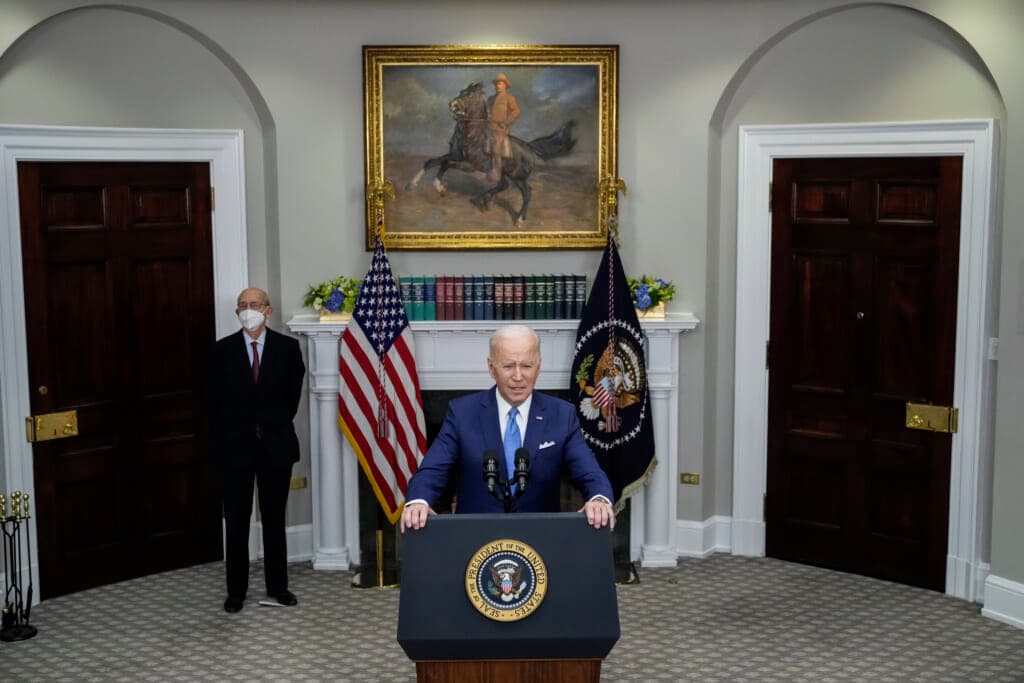 President Joe Biden stands behind a podium wearing a blue suit and light blue tie in front of a white fireplace