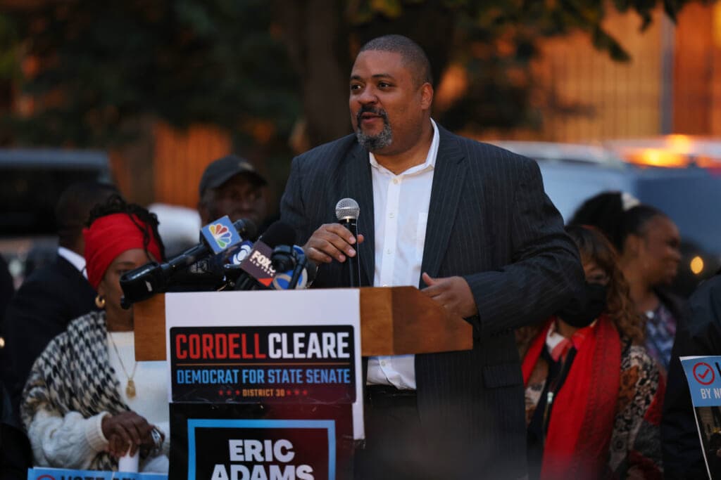 Alvin Bragg stands behind a podium with a microphone in his hand, wearing a gray jacket