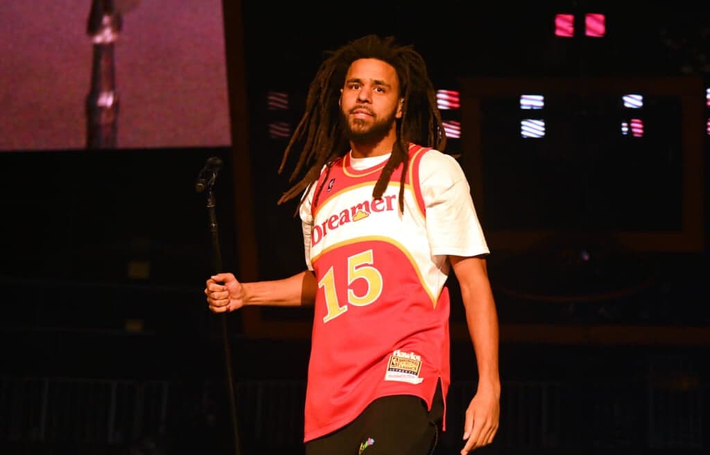 Hip-hop star J. Cole grasps a microphone stand wearing a red, white and yellow Number 15 Dreamer jersey