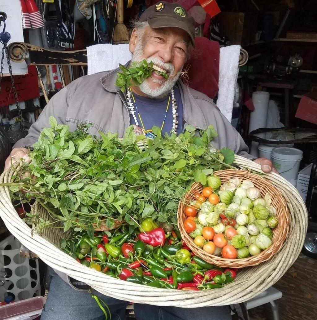 Daniel Chervoni with a plant sprig in his teeth and a large basket of produce in his lap, wearing a gray shirt