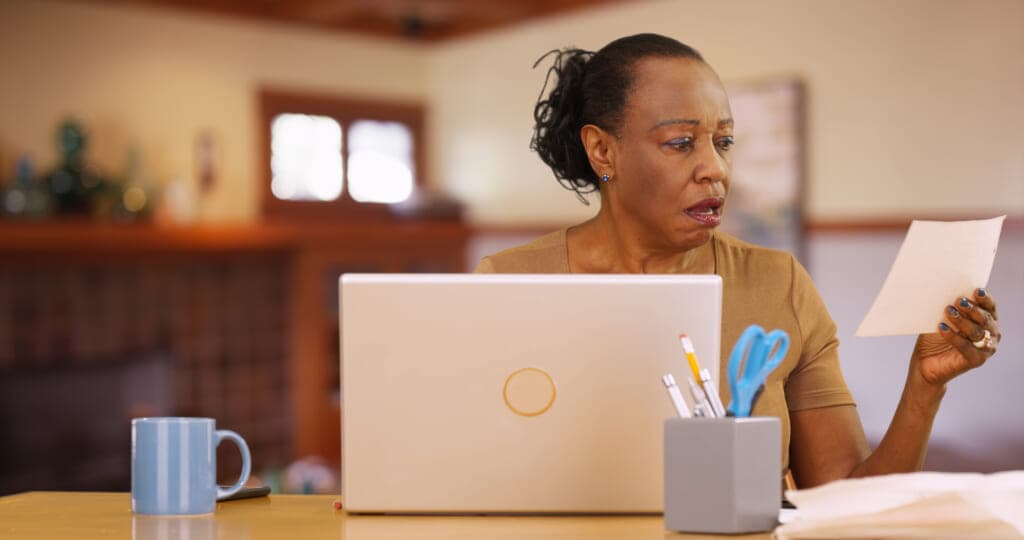 A woman at a laptop looking at a piece of paper