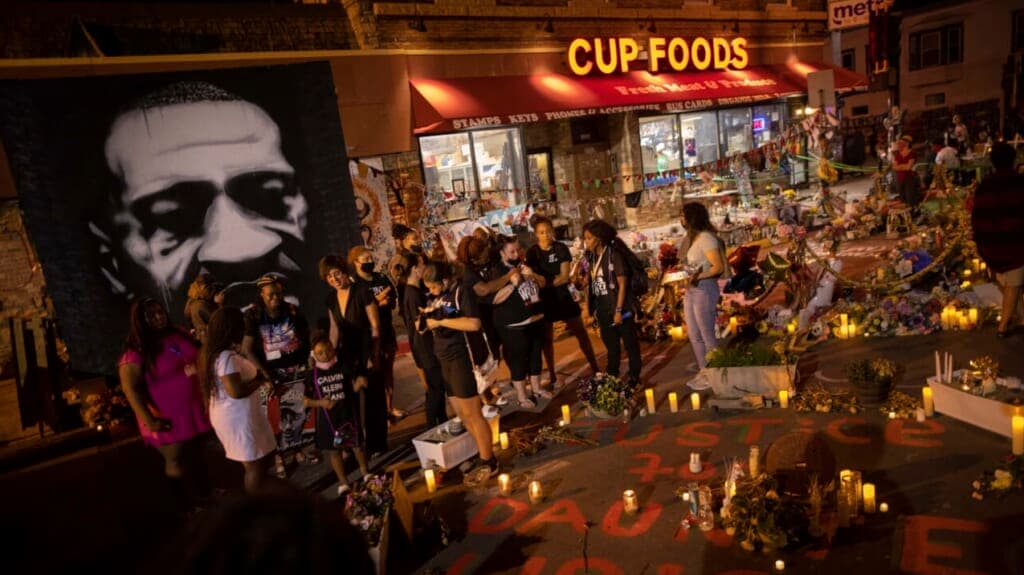 People gather in the parking lot of Cup Foods holding candles near a poster of George Floyd