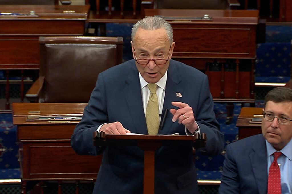 Senate Majority Leader Chuck Schumer of New York speaks behind a small wooden podium wearing a blue suit