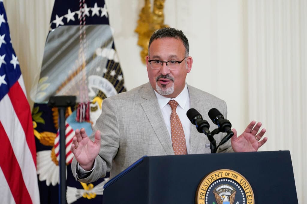 Education Secretary Miguel Cardona raises his hands behind a podium wearing a gray jacket and brown patterned tie