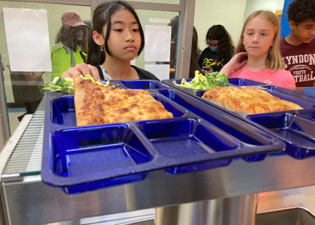 A girl reaches for a blue tray holding pizza and salad in a school cafeteria