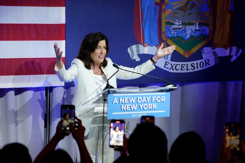 New York Gov. Kathy Hochul, wearing a white suit, raises her hands as she speaks at a podium