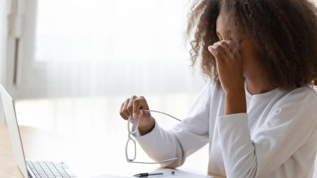 A woman in front of a laptop holding her head with eyeglasses in her hand