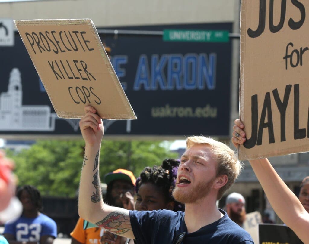 A tattooed blond man with a beard wearing a blue shirt holds a cardboard sign saying "Prosecute Killer Cops"