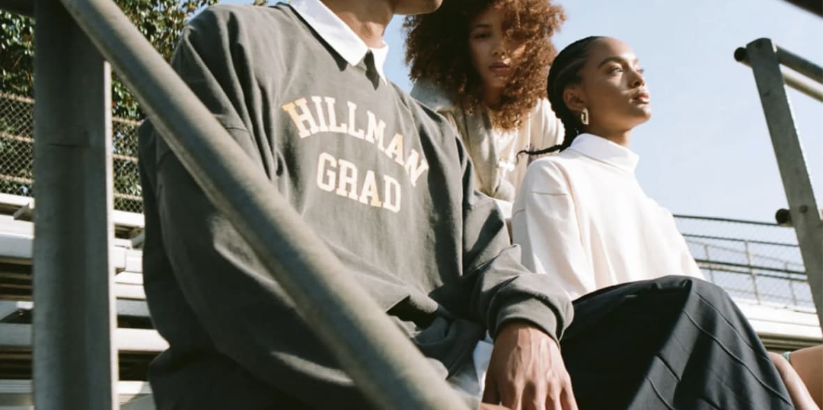 Three people sitting on bleachers outdoor, one wearing a "Hillman Grad" sweatshirt