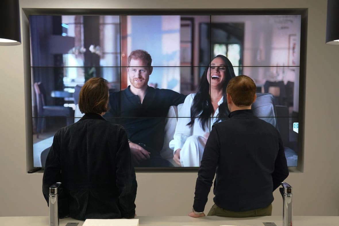 Two people watch a screen showing Prince Harry and Meghan Markle