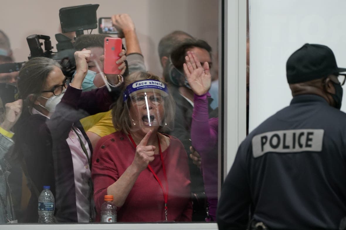 People wearing masks and holding cell phones yell as they look through a window where a police officer is standing