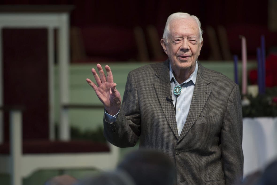 Former President Jimmy Carter waves, wearing a brown checked jacket and blue shirt