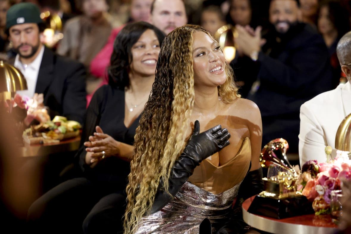 Singer Beyonce sits at a table in front of a Grammy award smiling while wearing a copper and silver gown