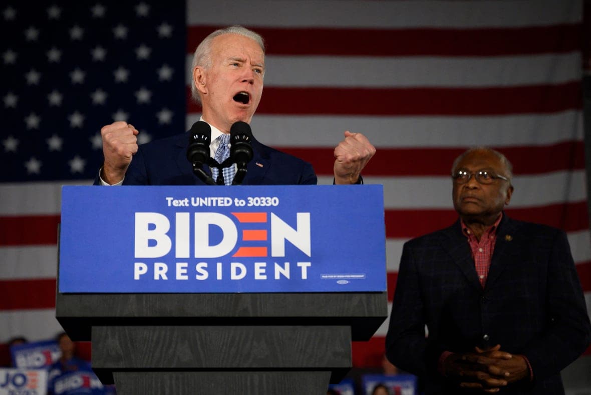 President Joe Biden gestures behind a podium reading "Biden President" wearing a dark suit and blue checked tie