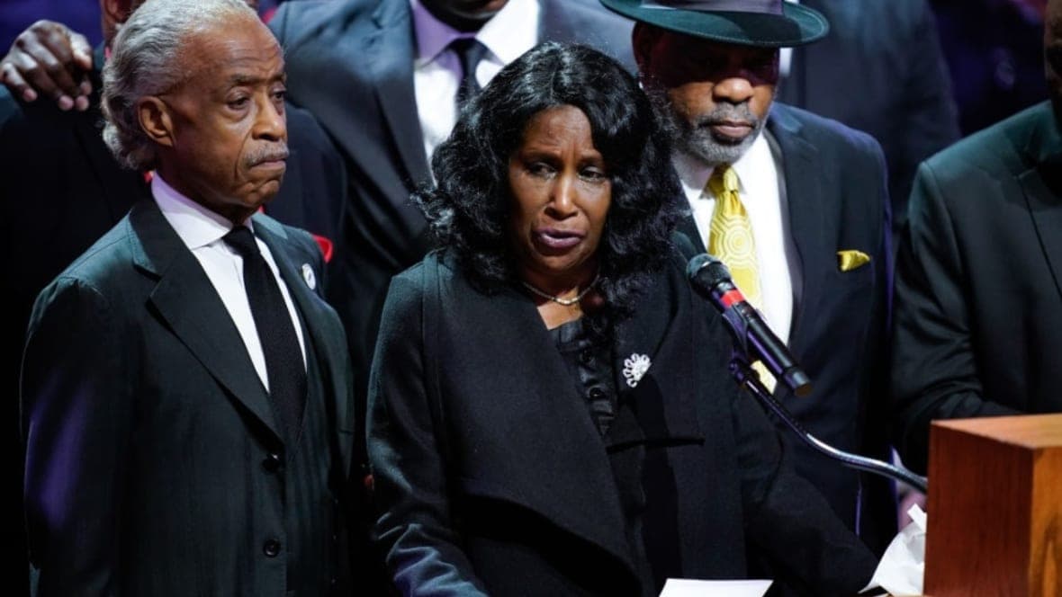 The Reverend Al Sharpton stands beside a woman speaking at a microphone, all wearing black