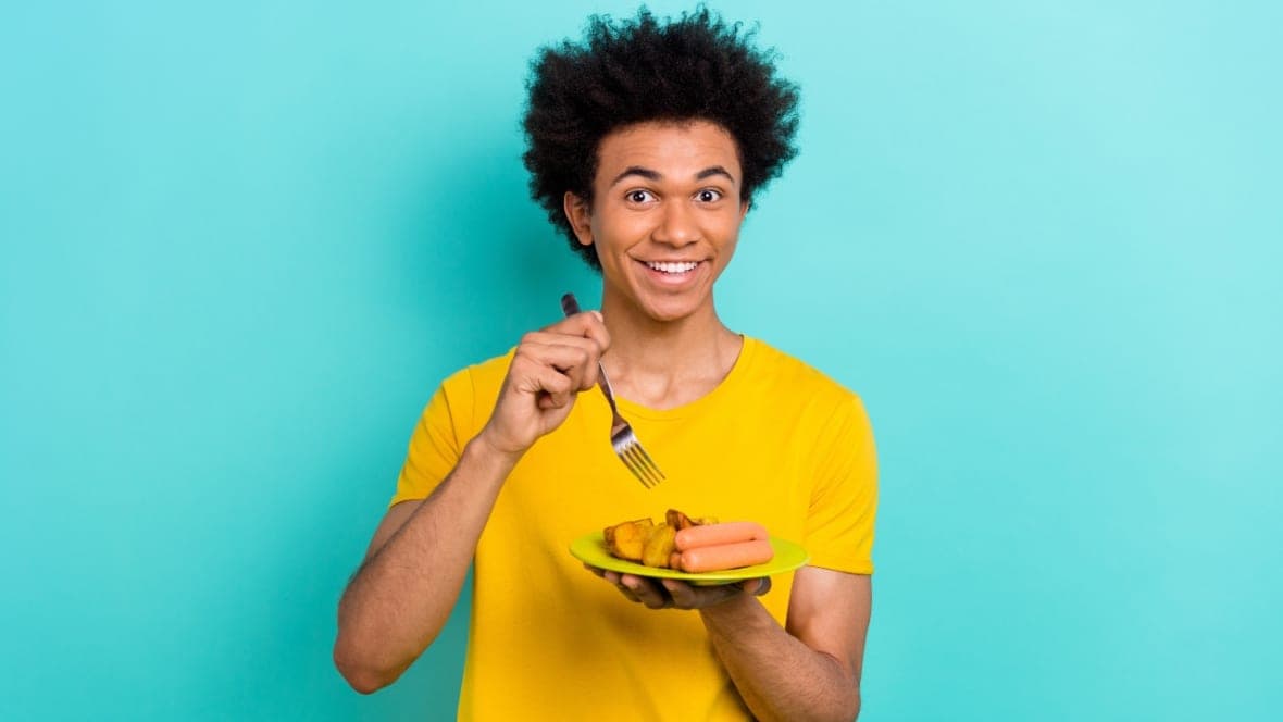 A man wearing a yellow shirt holds a silver fork over a yellow-green plate with hot dogs and other food