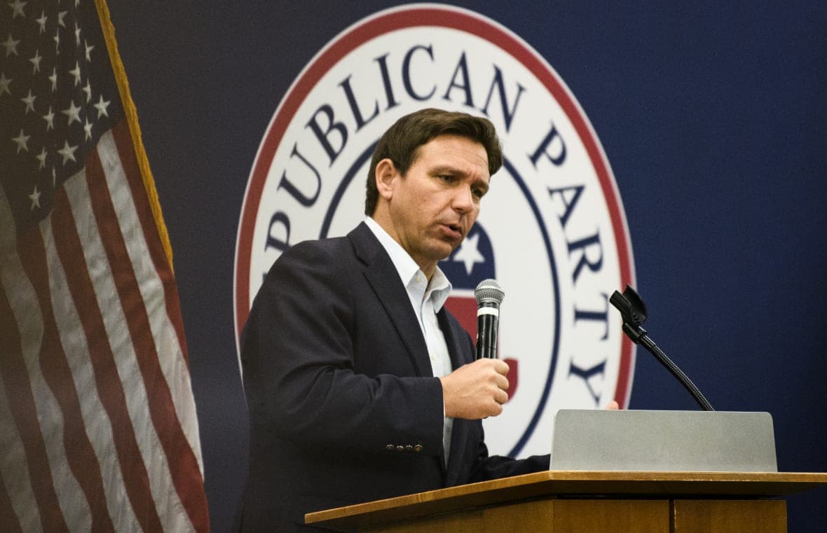 Republican Governor Ron DeSantis of Florida holds a microphone at a podium wearing a black jacket and white shirt