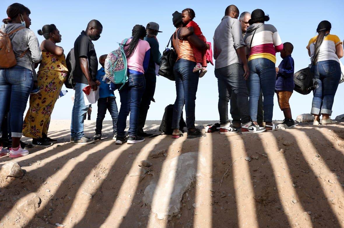 Haitian immigrants wait in line, shadowed by a fence