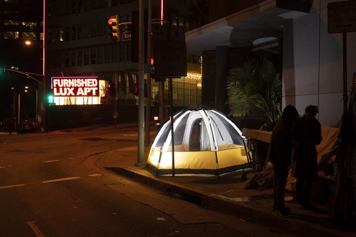 Several homeless people stand next to their tents across the street from a luxury apartment building
