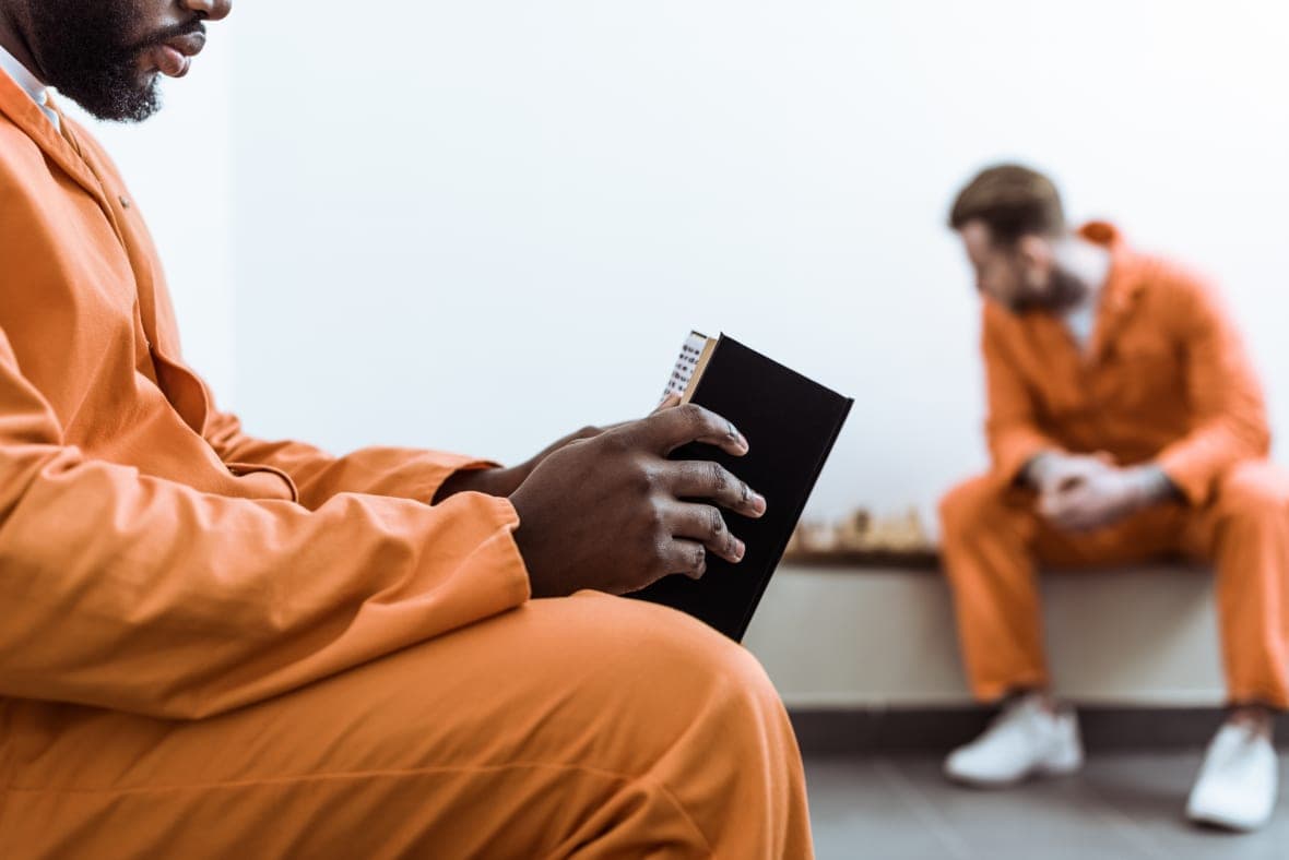 Two men in orange jumpsuits sitting, one reading a book