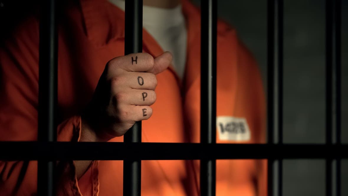A person behind bars in an orange jumpsuit with the word "Hope" tattooed on their knuckles
