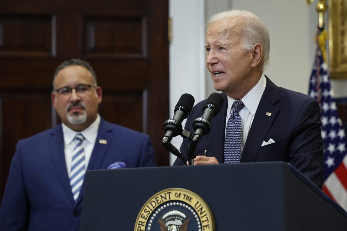 President Joe Biden stands at a podium with Education Secretary Miguel Cardona to his left, both wearing blue jackets
