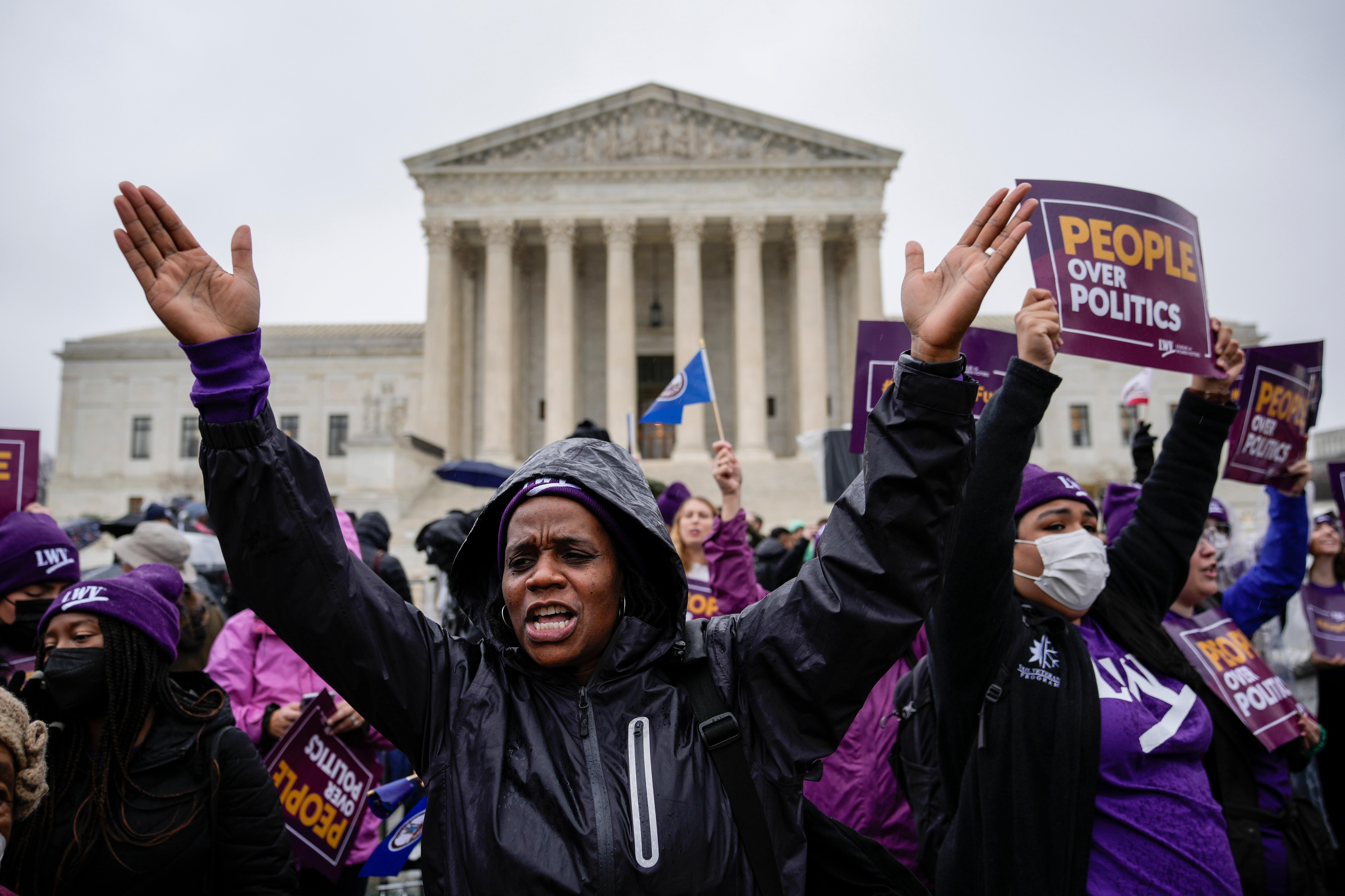 A woman wearing a black hooded jacket raises her hands in a crowd of people in front of the Supreme Court building