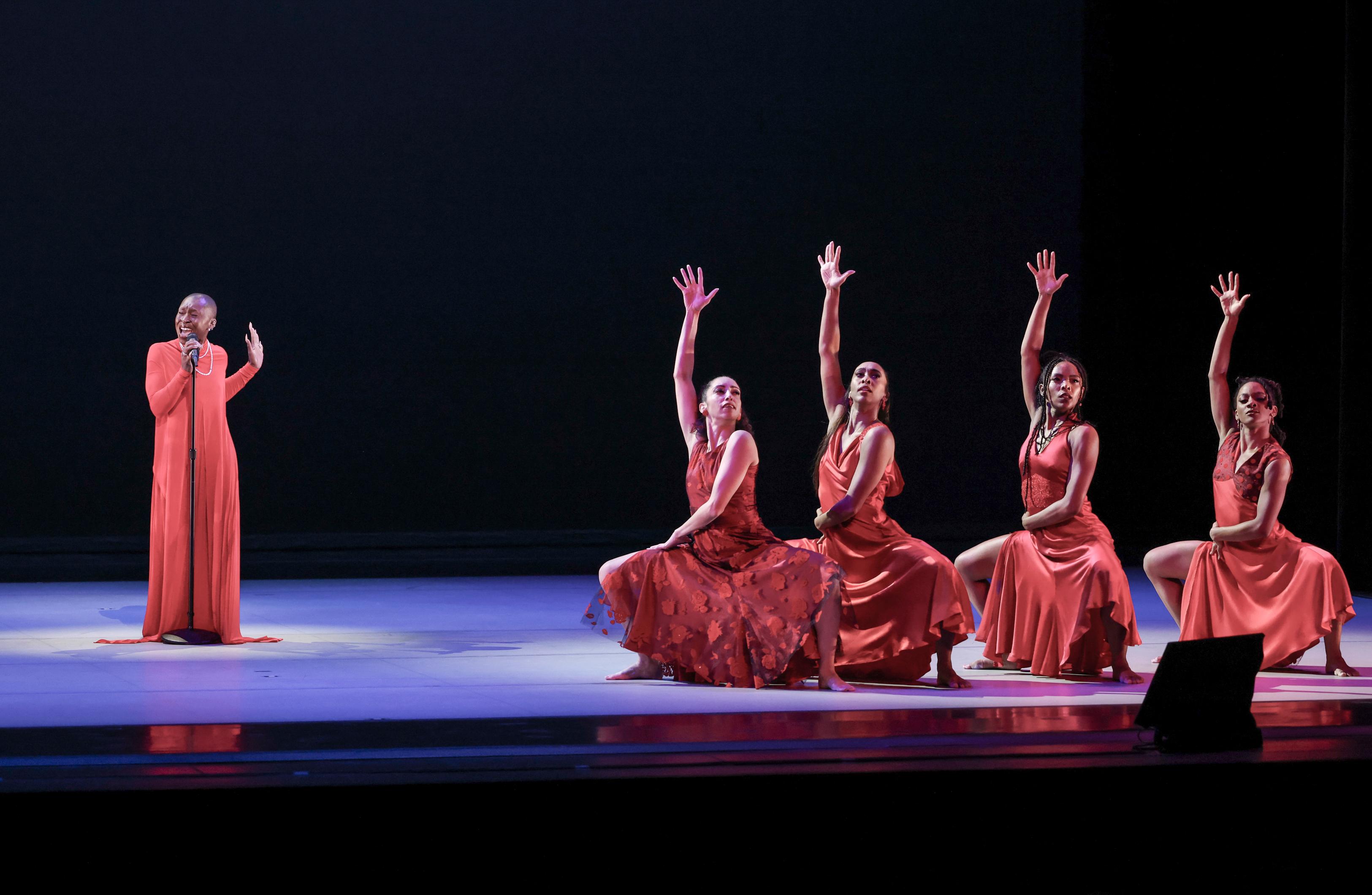 Singer Cynthia Erivo performs on stage wearing a long orange dress by four female dancers in shorter red dresses
