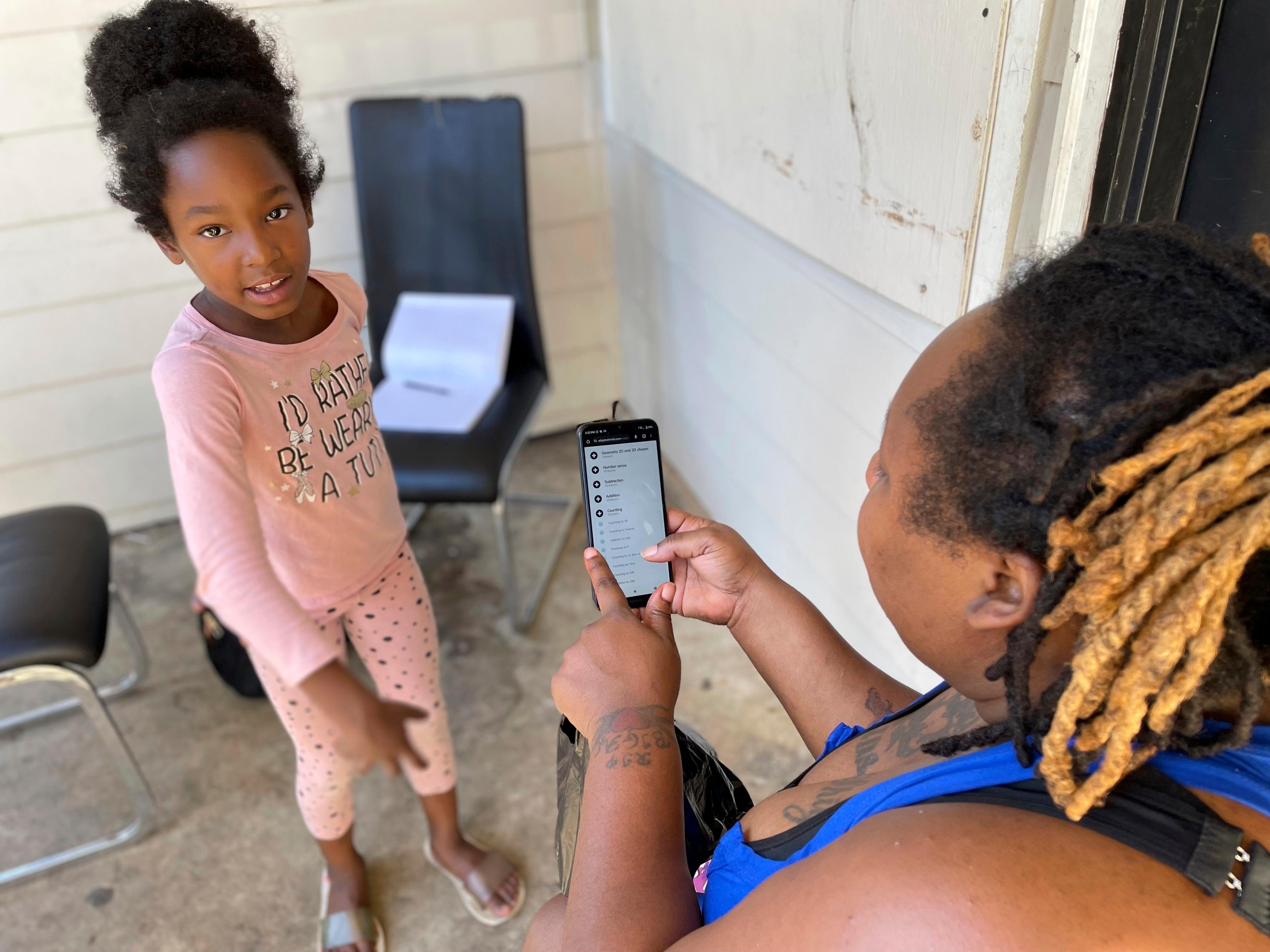 Tameka looks at a cell phone while her young daughter stands in front of her in pink pajamas