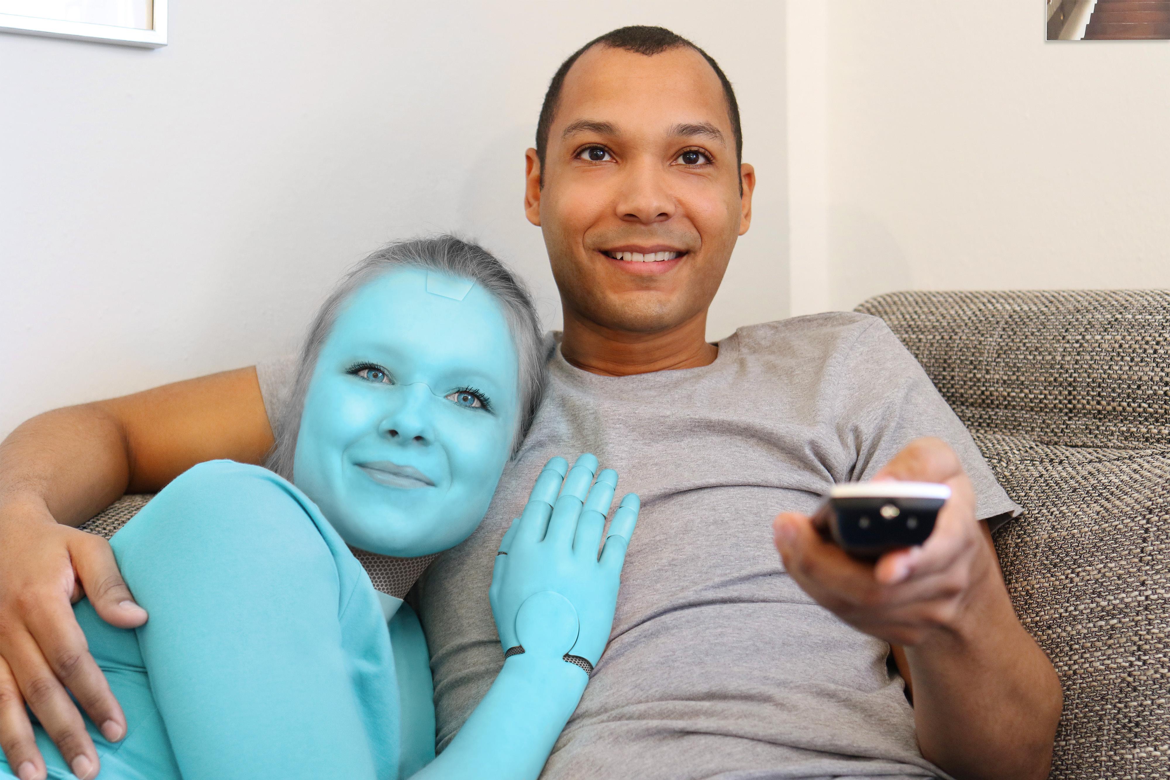 A man embraces a blue robotic woman on a couch