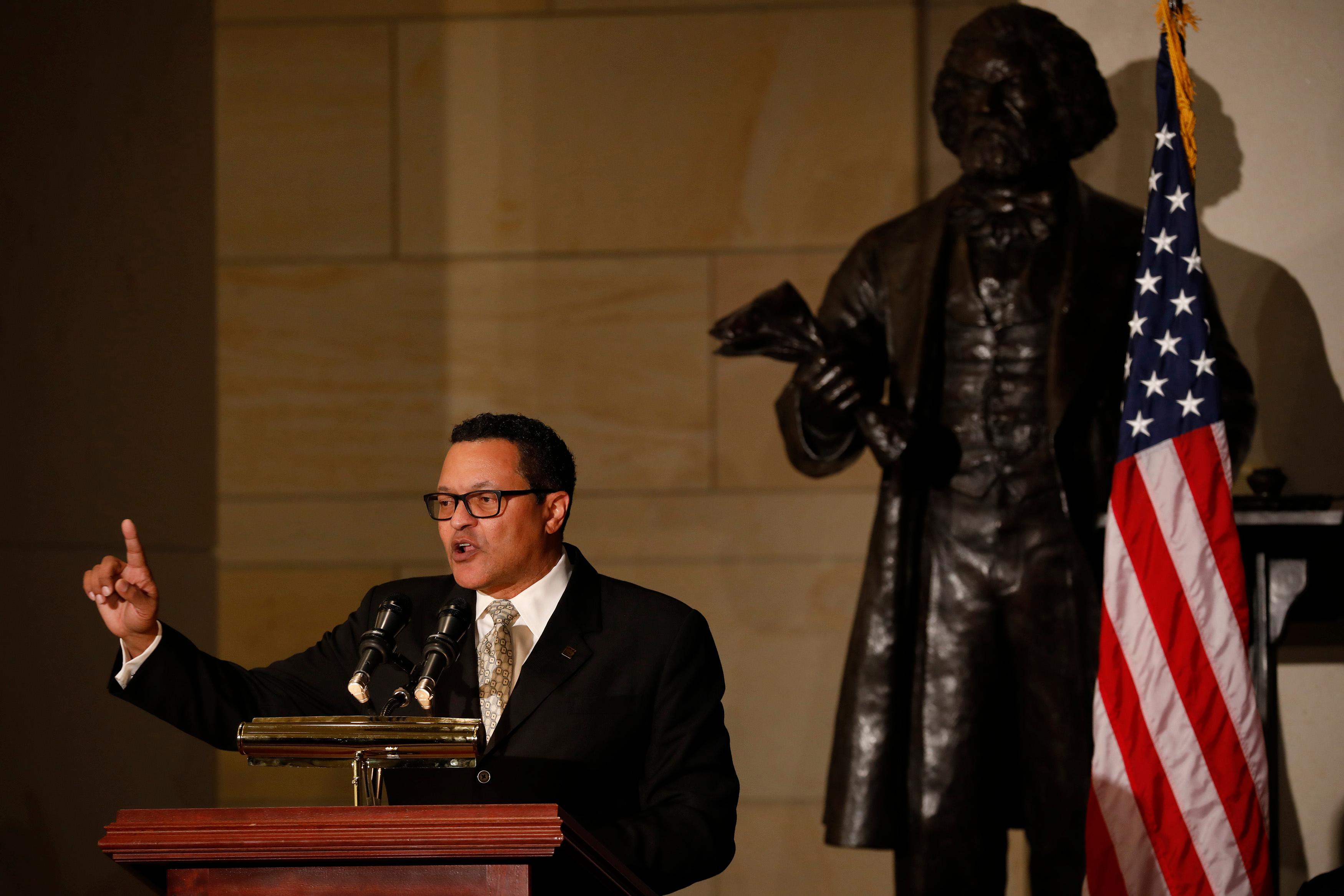 Ken Morris, a descendant of Frederick Douglass, speaks behind a podium wearing a black coat