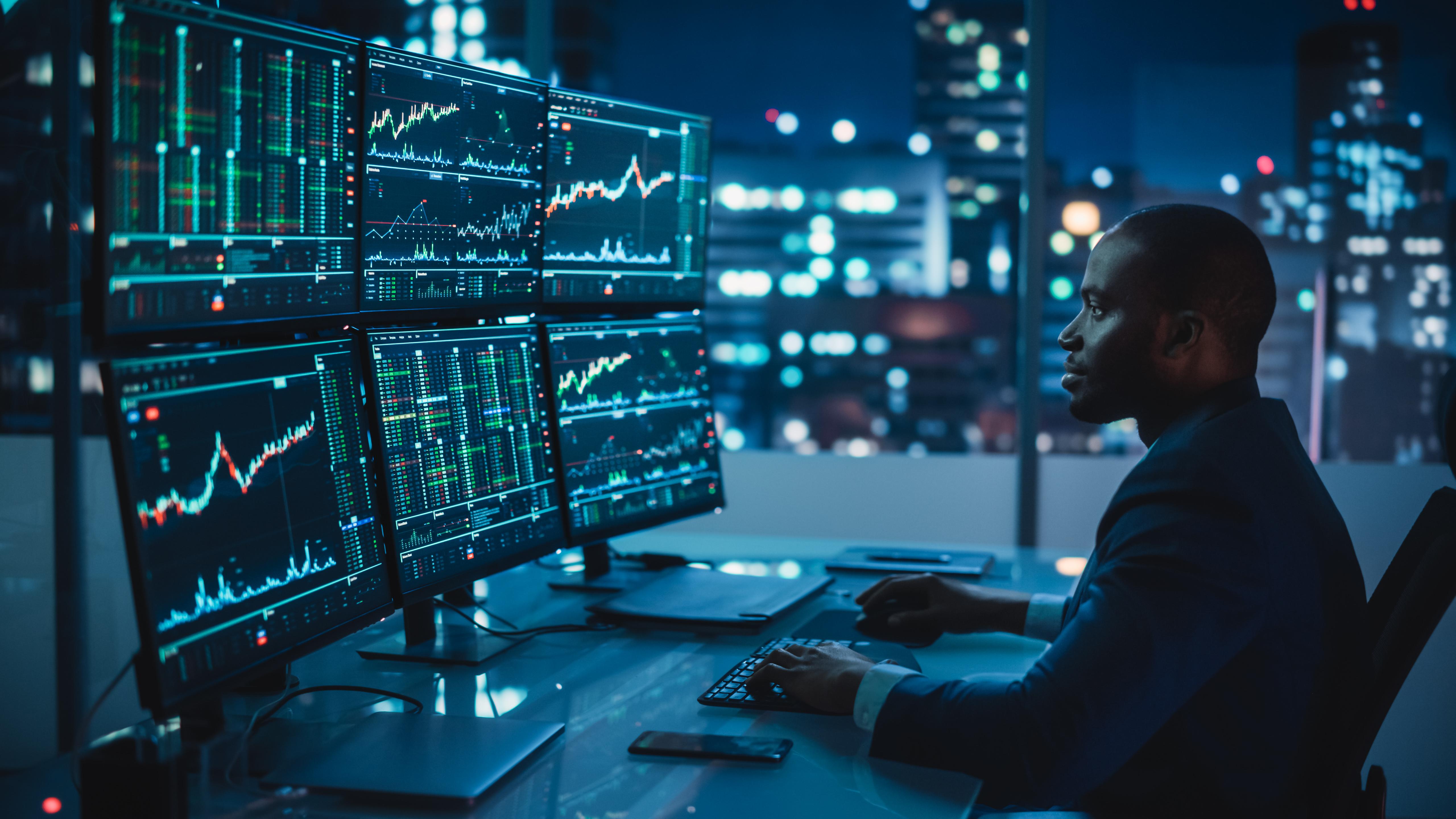 A man sitting at a desk with 6 computer screens showing graphs and information