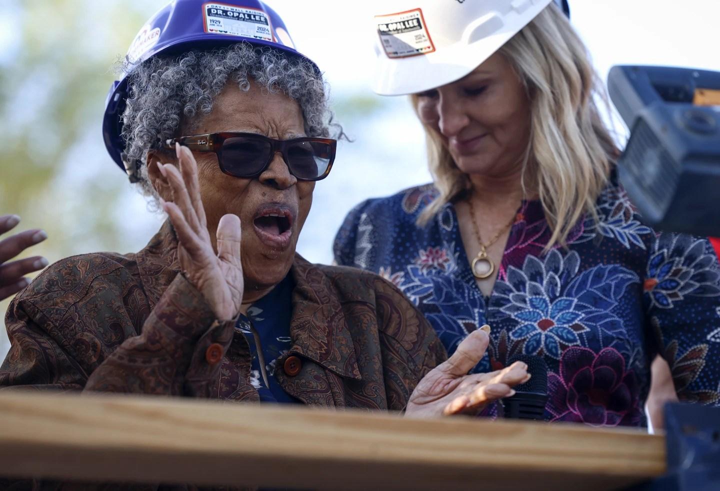 Opal Lee, wearing a purple hard hat and brown coat, applauds during a ceremony to build her new home