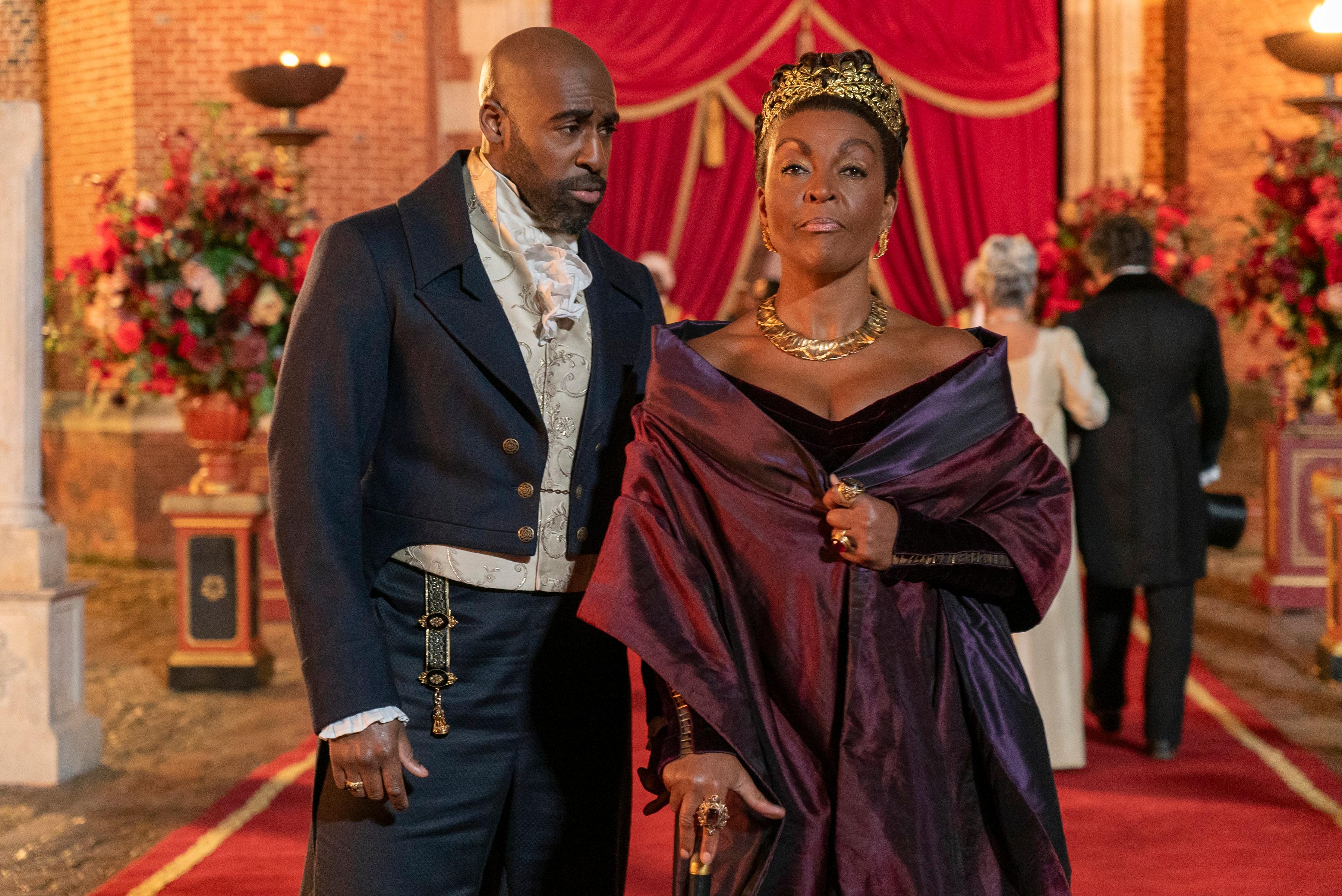 Actors Daniel Francis in a formal suit and Adjoa Andoh wearing a black gown, purple wrap and tiara in "Bridgerton"