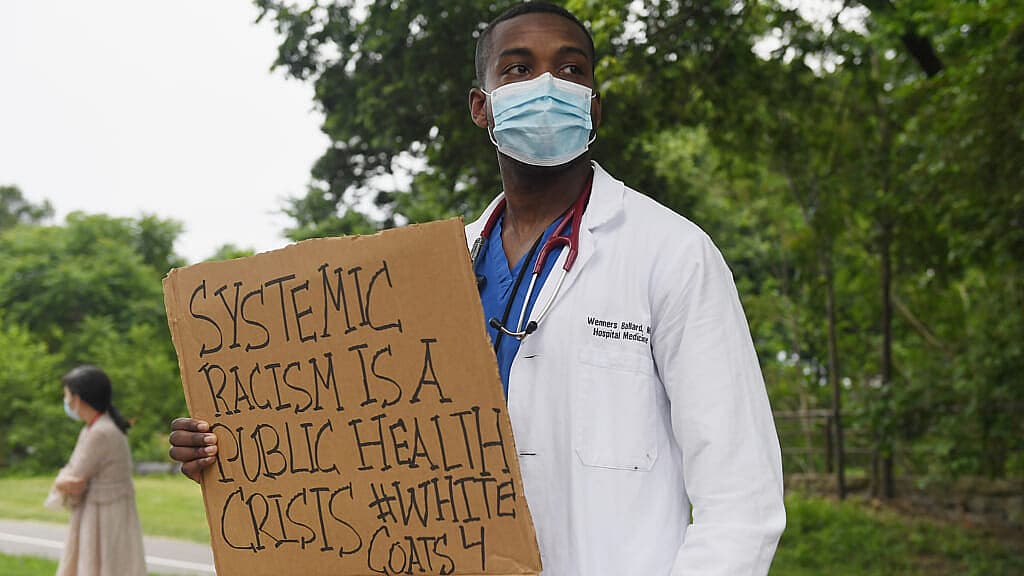 A man wearing a doctor's coat and blue scrubs holds a sign reading "Systemic racism is a public health crisis"