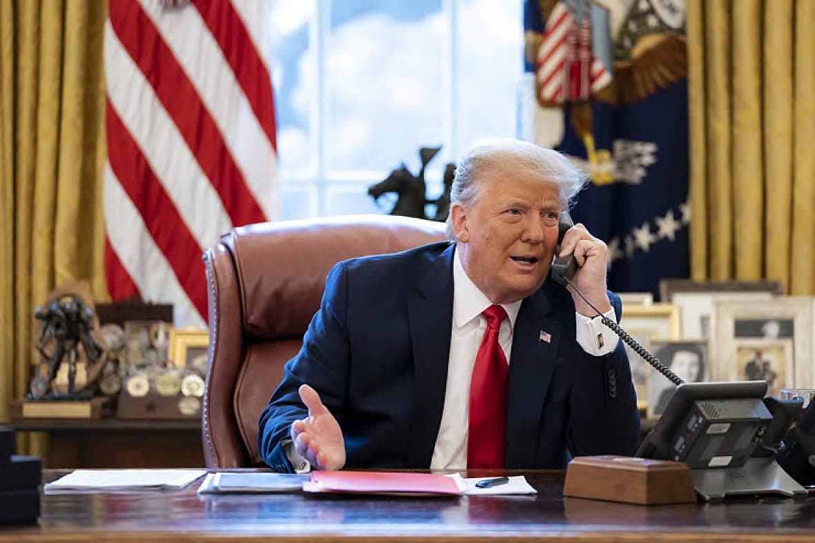 Former President Donald Trump speaks on a corded phone while sitting at a desk, wearing a blue jacket and red tie