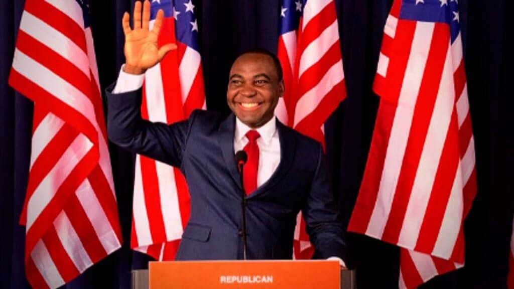 Republican Michigan governor candidate Austin Chenge waving from behind a podium, wearing a blue suit and red tie