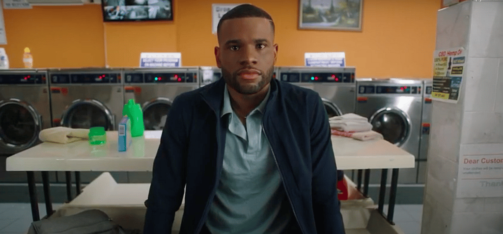 Actor Reggie Lochard stands before a table in a laundromat wearing a navy jacket and light green shirt