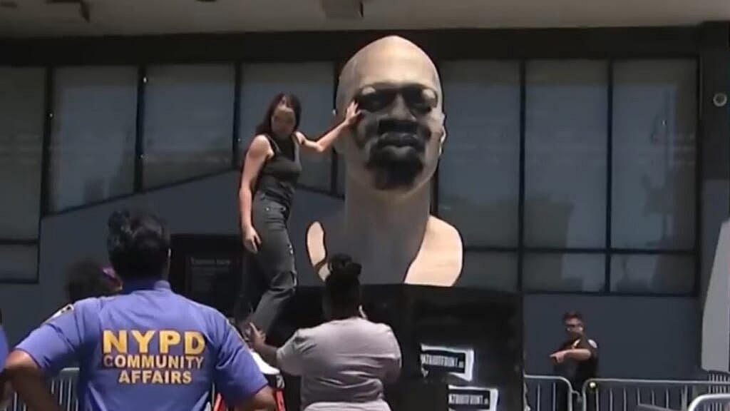 A woman in a black tank top and pants stands before a statue of George Floyd defaced with black paint