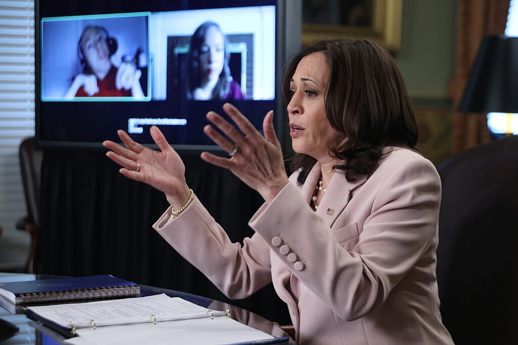 Vice President Kamala Harris gestures as she sits at a desk, wearing a pale pink jacket