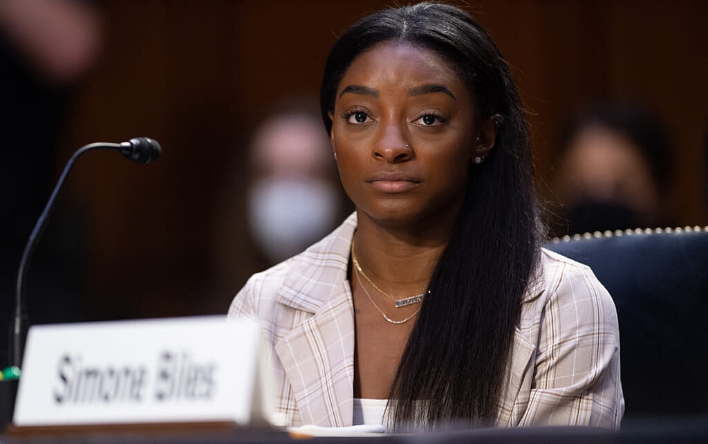 Olympic gymnast Simone Biles sits at a table wearing a plaid jacket