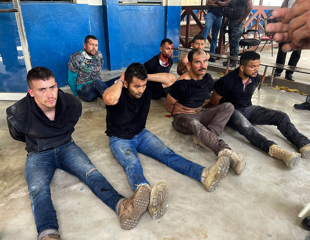Seven men in handcuffs sit on a floor